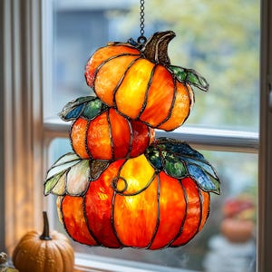 Könnte beinhalten: Ein Buntglas-Suncatcher mit drei gestapelten Kürbissen in leuchtenden Orange-, Gelb- und Rottönen. Die Kürbisse sind mit grünen und blauen Glasblättern akzentuiert, die durch dunkle Metalllötstellen und eine Aufhängungskette verbunden sind.