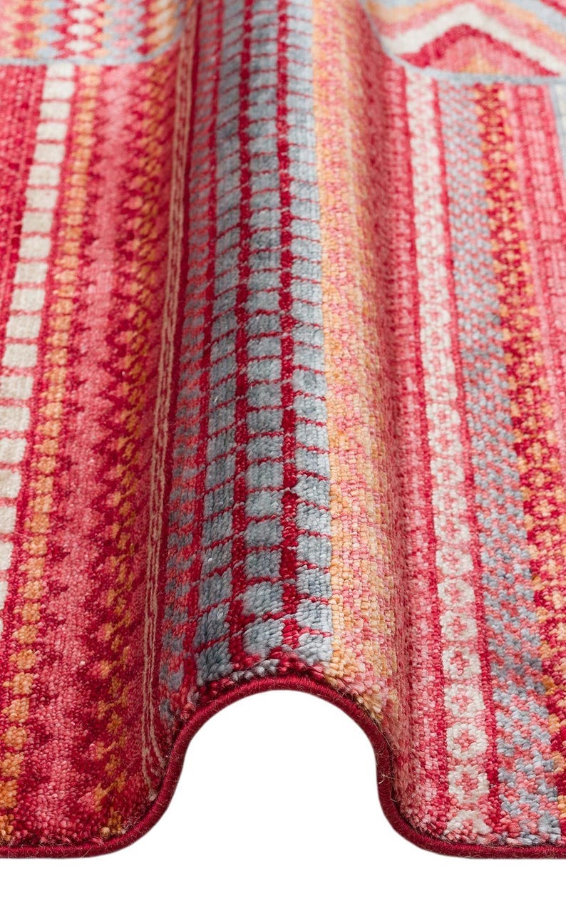 May include: A textile rug featuring a vibrant pattern of vertical stripes in shades of red, pink, orange, and light blue. The surface displays a variety of woven textures, including grid-like squares, horizontal bands, and intricate geometric motifs.
