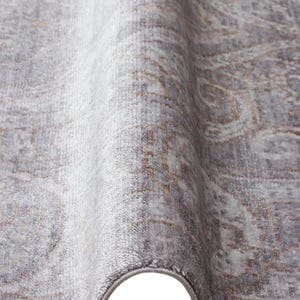 May include: A rectangular area rug featuring a soft, low-pile texture in shades of muted gray and light brown. The surface displays a faded, intricate damask pattern with swirling motifs and a finished, bound edge in a matching gray tone.