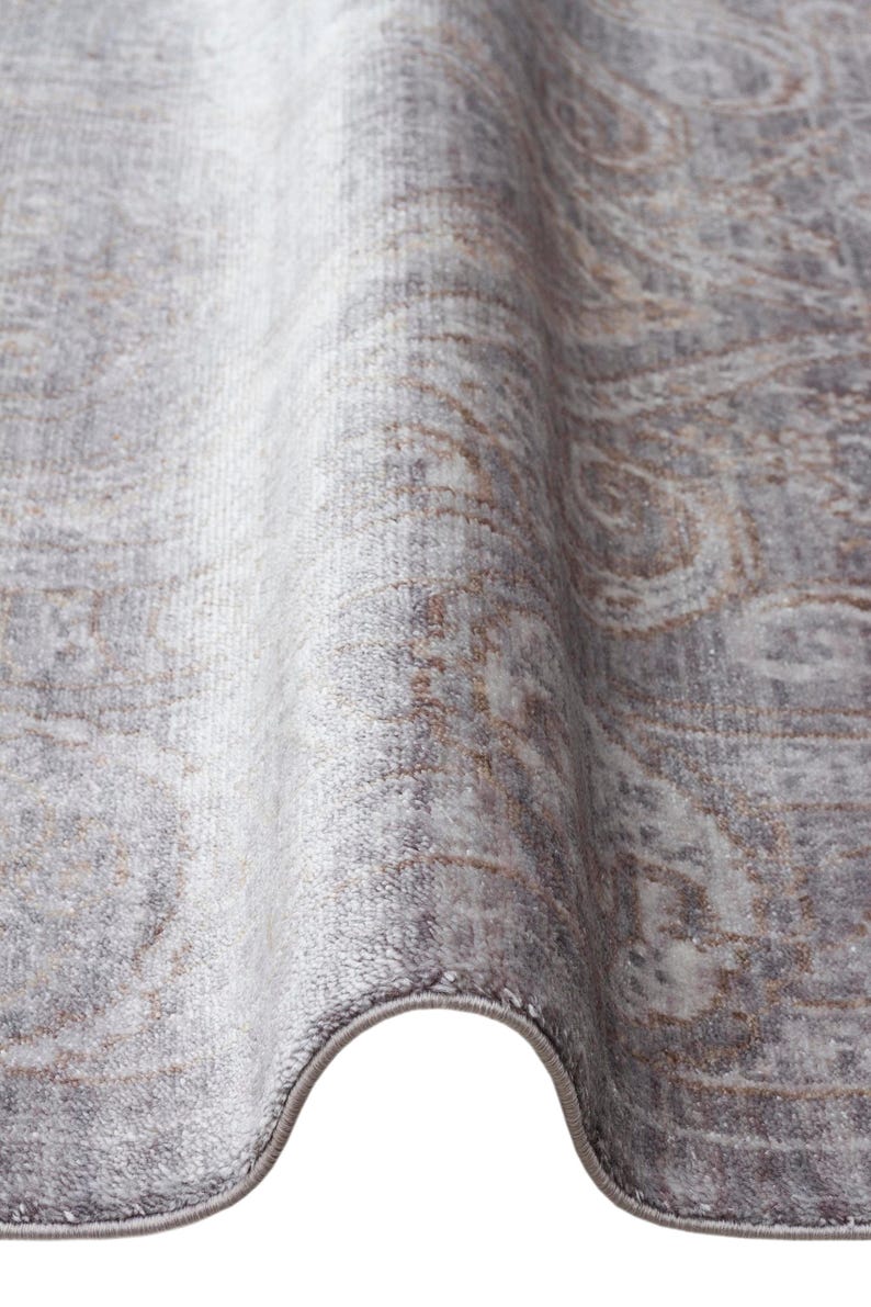 May include: A rectangular area rug featuring a soft, low-pile texture in shades of muted gray and light brown. The surface displays a faded, intricate damask pattern with swirling motifs and a finished, bound edge in a matching gray tone.