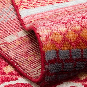 May include: A woven area rug featuring a vibrant red, orange, and light blue geometric pattern. The rug is folded over to reveal a dense, textured pile and a finished red edge binding, showcasing the intricate weave and color palette of the textile.
