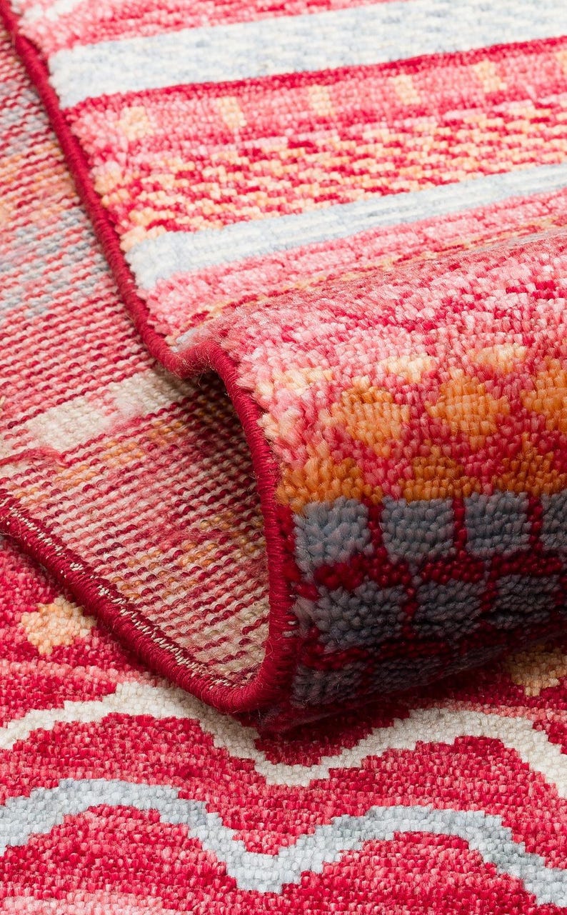 May include: A woven area rug featuring a vibrant red, orange, and light blue geometric pattern. The rug is folded over to reveal a dense, textured pile and a finished red edge binding, showcasing the intricate weave and color palette of the textile.