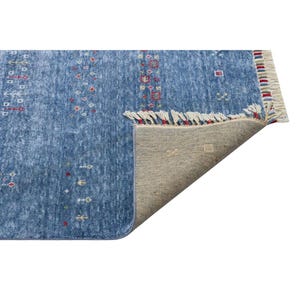 May include: A blue area rug with a mottled texture and small, repeating geometric patterns in red, yellow, and white. The rug features a folded corner revealing a beige woven backing and a short fringe edge with multicolored thread accents.