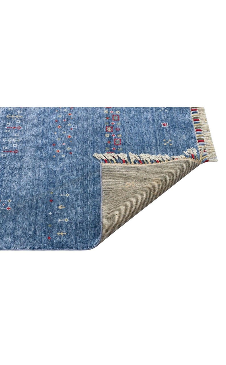 May include: A blue area rug with a mottled texture and small, repeating geometric patterns in red, yellow, and white. The rug features a folded corner revealing a beige woven backing and a short fringe edge with multicolored thread accents.