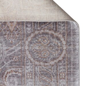 May include: A rectangular area rug featuring a muted gray and beige color palette with a repeating medallion and floral pattern. The corner of the rug is folded over to reveal a light beige, textured woven backing and a finished bound edge.
