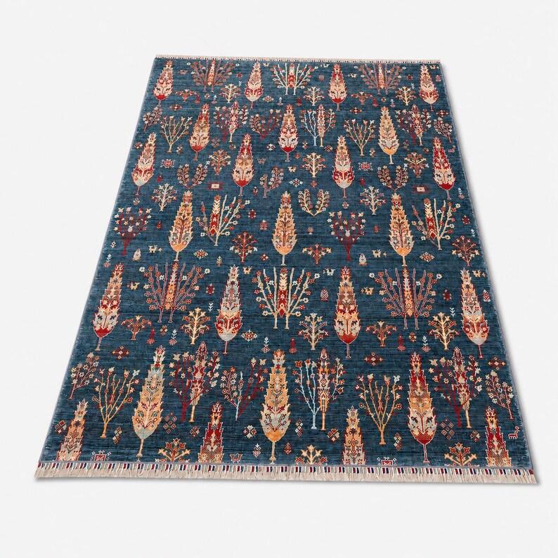 May include: A rectangular area rug featuring a deep blue background with a repeating pattern of stylized trees and floral motifs in shades of red, orange, cream, and light blue. The rug has a short fringe along the top and bottom edges.