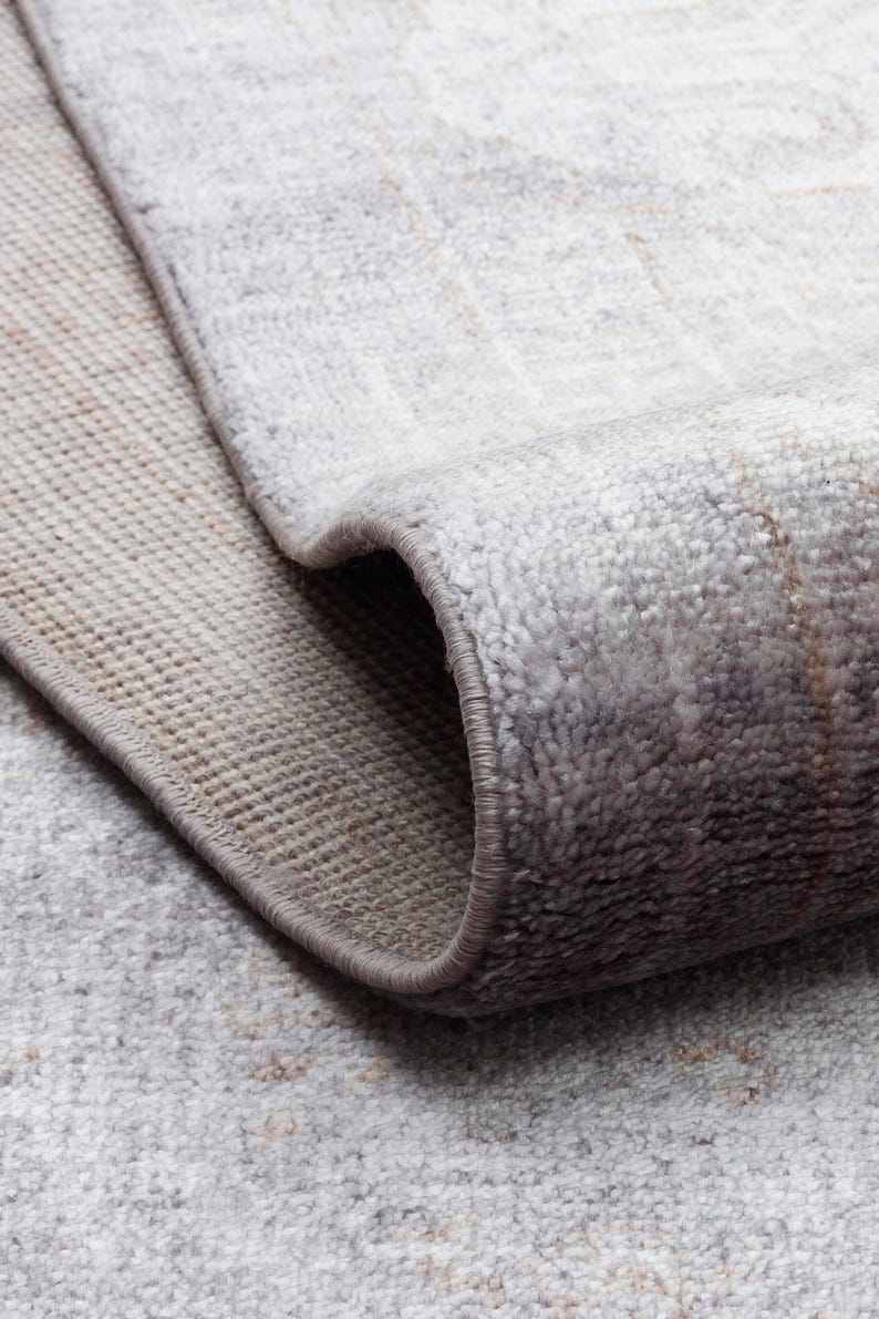 May include: A rolled area rug featuring a light gray and beige color palette with a faded, distressed pattern. The rug has a soft, textured pile surface and a finished, stitched edge that follows the curve of the roll, revealing the backing.