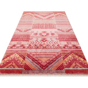 May include: A rectangular area rug featuring a vibrant red, orange, and pink color palette with intricate geometric patterns including chevrons, horizontal stripes, and diamond shapes. The rug has a short fringe along the bottom edge.