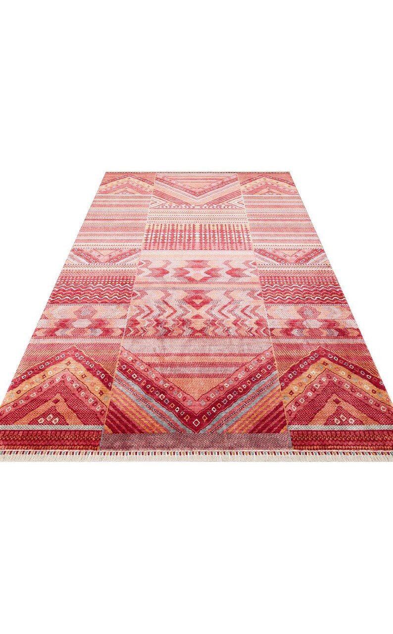 May include: A rectangular area rug featuring a vibrant red, orange, and pink color palette with intricate geometric patterns including chevrons, horizontal stripes, and diamond shapes. The rug has a short fringe along the bottom edge.