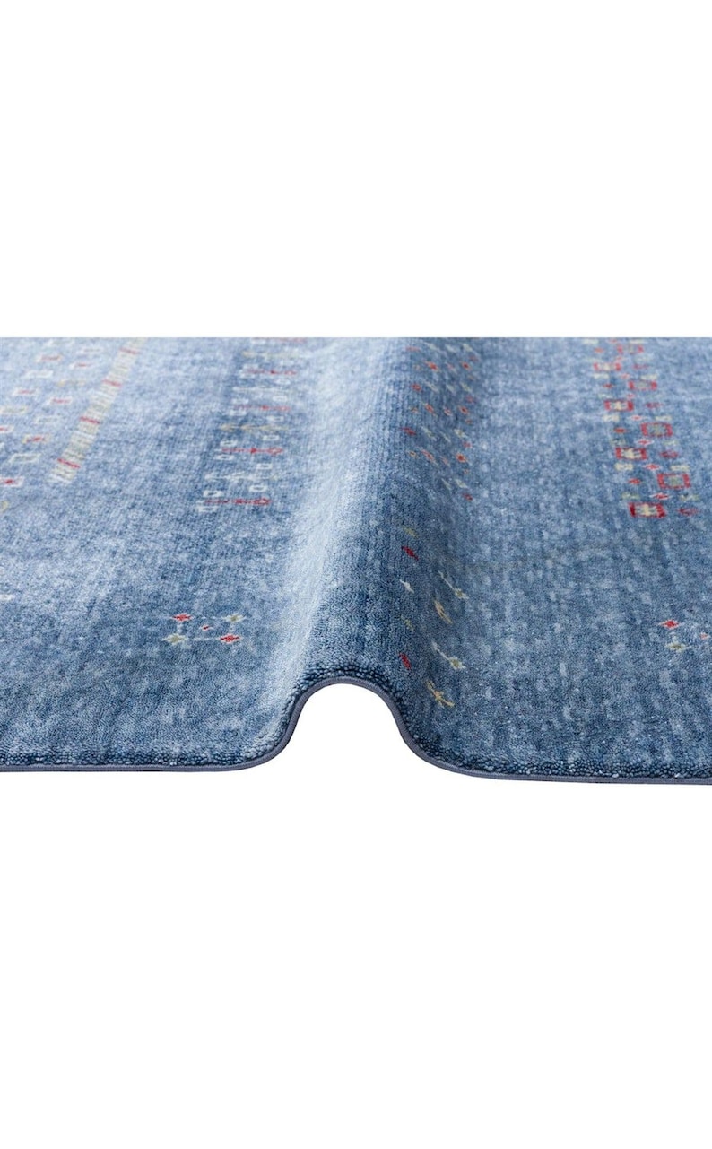 May include: A blue area rug with a textured, soft pile surface featuring a repeating pattern of small, scattered geometric shapes in red, white, and yellow. The rug is folded over to reveal its thickness and the finished, bound edge along the bottom.