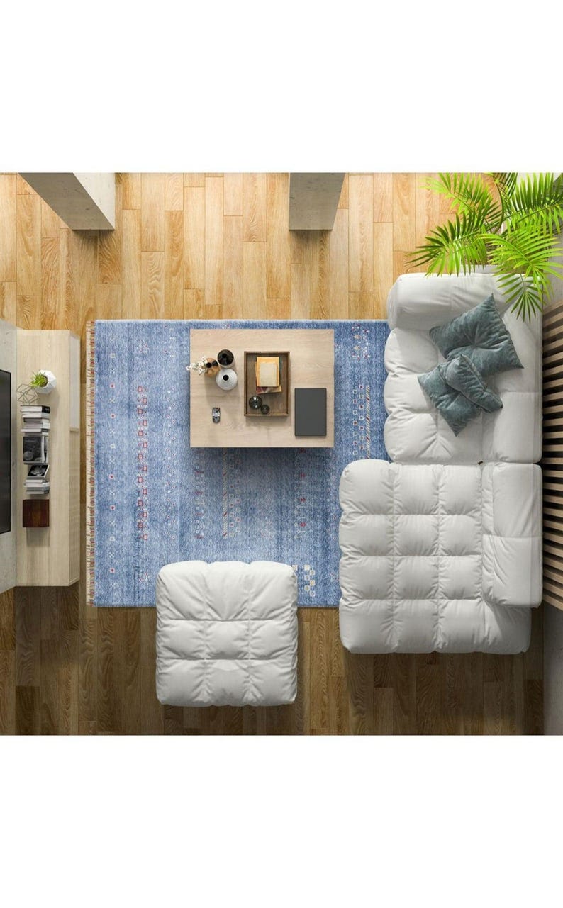 May include: A white tufted sectional sofa with a matching ottoman, positioned on a blue patterned area rug. A light wood coffee table sits in the center, holding a tray with small decorative items, a book, and a black rectangular object.