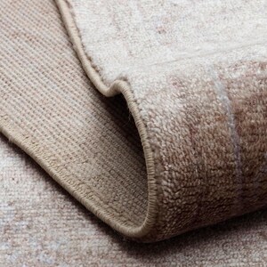 May include: A beige and cream area rug with a soft, textured pile and a folded corner revealing the underside. The rug features a subtle, faded pattern in neutral tones with a finished, stitched edge that runs along the perimeter of the fabric.