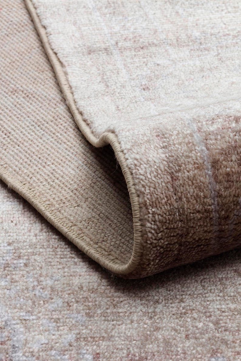 May include: A beige and cream area rug with a soft, textured pile and a folded corner revealing the underside. The rug features a subtle, faded pattern in neutral tones with a finished, stitched edge that runs along the perimeter of the fabric.