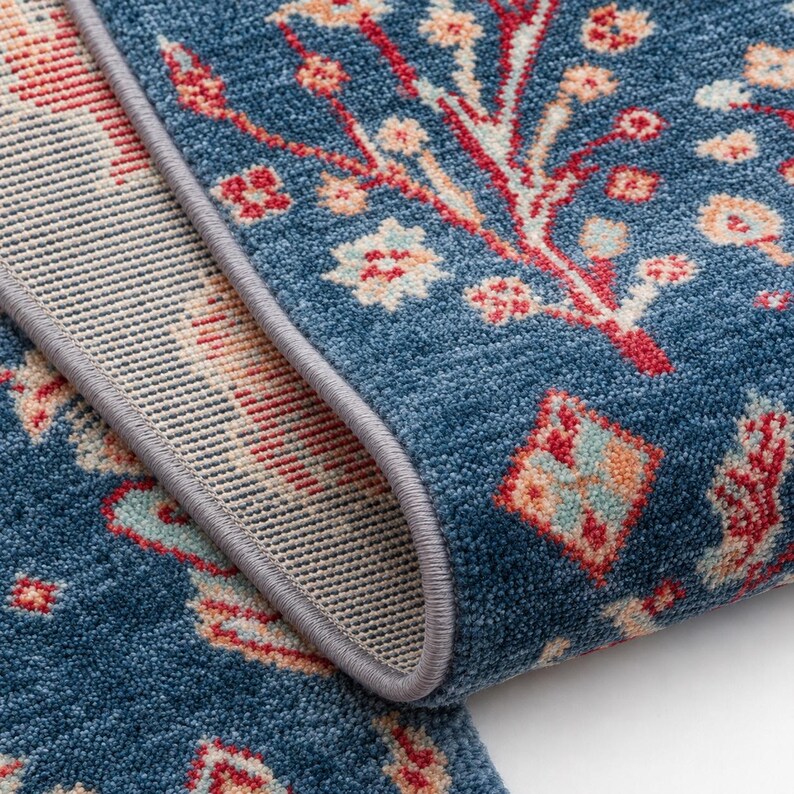 May include: A blue area rug featuring a repeating floral pattern in shades of red, cream, and light blue. The rug has a thick, textured pile and is finished with a grey serged edge that highlights the construction of the fabric and the backing.