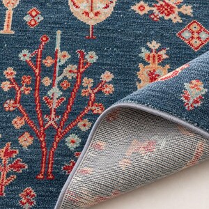 May include: A deep blue area rug featuring a repeating pattern of stylized trees and geometric floral motifs in shades of red, cream, and light blue. The rug has a textured pile surface and a gray bound edge, with the underside partially folded back.