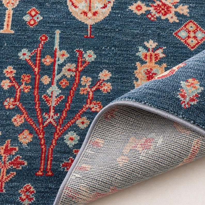 May include: A deep blue area rug featuring a repeating pattern of stylized trees and geometric floral motifs in shades of red, cream, and light blue. The rug has a textured pile surface and a gray bound edge, with the underside partially folded back.