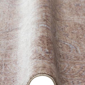 May include: A beige and light brown area rug featuring a faded, intricate pattern with soft, muted tones. The rug is shown folded, revealing its textured surface, dense pile, and a finished, bound edge that runs along the curved bottom border.