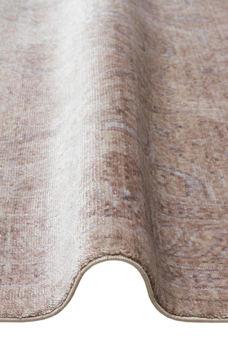 May include: A beige and light brown area rug featuring a faded, intricate pattern with soft, muted tones. The rug is shown folded, revealing its textured surface, dense pile, and a finished, bound edge that runs along the curved bottom border.