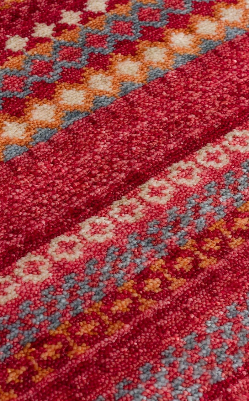 May include: A textured rug featuring a vibrant red base with horizontal bands of intricate geometric patterns. The design incorporates repeating diamond and floral motifs in shades of cream, soft orange, and muted blue, creating a dense pile.