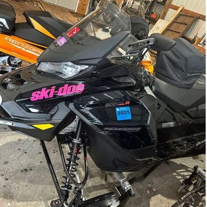 Puede incluir: Una moto de nieve Ski-Doo Renegade 900R negra con letras rosas. La moto de nieve tiene un asiento negro y un parabrisas. La moto de nieve est&aacute; sobre una superficie de hormig&oacute;n.