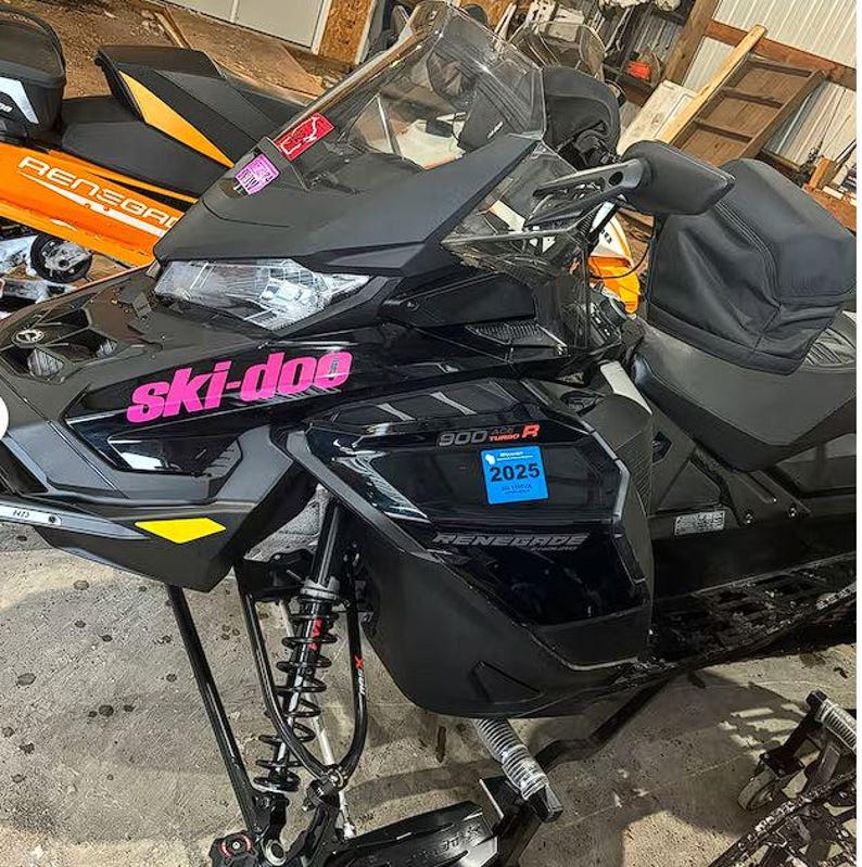 Puede incluir: Una moto de nieve Ski-Doo Renegade 900R negra con letras rosas. La moto de nieve tiene un asiento negro y un parabrisas. La moto de nieve est&aacute; sobre una superficie de hormig&oacute;n.