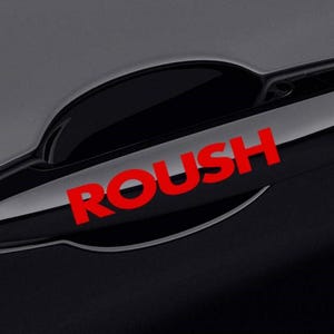 Puede incluir: Un tirador de puerta de coche negro con la palabra "ROUSH" en rojo impresa. El tirador es brillante y refleja el entorno. El tirador de la puerta del coche es un accesorio para el coche.
