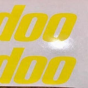 Puede incluir: Cuatro calcoman&iacute;as de vinilo amarillo con el texto "ski-doo" impreso en ellas. Las calcoman&iacute;as est&aacute;n sobre un fondo blanco.
