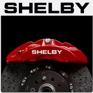 May include: A red Shelby brake caliper with the Shelby logo in white, mounted on a black rotor. The word "SHELBY" is also displayed in white at the top of the image against a white background.