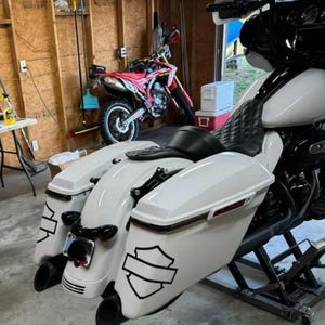 May include: A white motorcycle with a black diamond-stitched seat and dual saddlebags featuring black shield-shaped decals. The motorcycle is positioned on a metal lift, showcasing its rear fender, dual exhaust pipes, and black trim details.