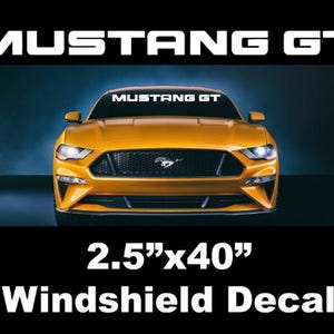 May include: Mustang GT windshield decal, 2.5" x 40", in white text. The image shows a yellow Mustang GT car. The decal is designed to fit across the top of the windshield. The car has a black grill and headlights.