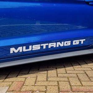 May include: A section of a bright blue car door with the white text "MUSTANG GT" in a bold, sans-serif font. The car's body is glossy, and the image is taken outdoors.