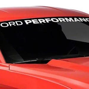 May include: A red car with a black windscreen. The words "FORD PERFORMANCE" are displayed in white across the top of the windscreen. The car's bonnet is visible, and the image is well-lit.