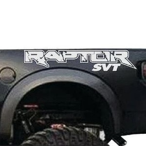 May include: Black truck bed with white "RAPTOR SVT" decal. The decal is stylized with a bold font and a sharp, angular design. The truck bed features a wheel well, taillight, and a portion of the tire.