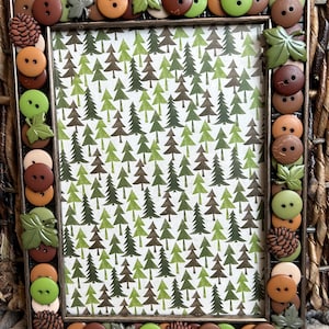 May include: A brown picture frame decorated with various buttons, including green, brown, and beige buttons. The buttons are arranged in a pattern around the frame. The frame has a white background with a pattern of green and brown trees.