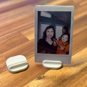 May include: White plastic photo stand with a Polaroid photo of a woman and two children. The photo shows a woman with brown hair and a light blue shirt, a young boy with brown hair and an orange shirt, and a girl with brown hair and a blue shirt. The photo stand is on a wooden surface.