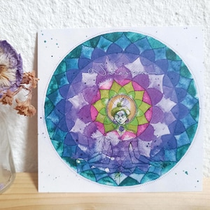 Pode incluir: Uma impressão quadrada em aquarela com uma figura meditando dentro de um mandala vibrante. O mandala apresenta um espectro de cores, incluindo azuis, roxos, verdes e rosas, com a palavra "Sahasrara" inscrita. A obra de arte está sobre um fundo branco.