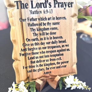 May include: A wooden plaque with the Lord's Prayer engraved on it. The plaque is in the shape of a scroll and has a brown wooden stand. The text on the plaque reads: "The Lord's Prayer, Matthew 6:9-13, Our Father which art in heaven, Hallowed be thy name, Thy kingdom come, Thy will be done, On earth, as it is in heaven. Give us this day our daily bread. And forgive us our trespasses, as we forgive those who trespass against us. Lead us not into temptation, But deliver us from evil. For thine is the kingdom, the power and the glory, for ever and ever, Amen."