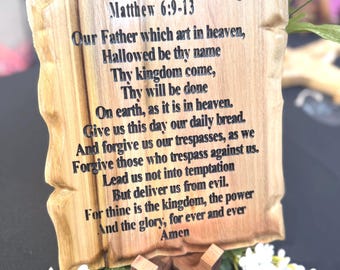 May include: A wooden plaque with the Lord's Prayer engraved on it. The plaque is in the shape of a scroll and has a brown wooden stand. The text on the plaque reads: "The Lord's Prayer, Matthew 6:9-13, Our Father which art in heaven, Hallowed be thy name, Thy kingdom come, Thy will be done, On earth, as it is in heaven. Give us this day our daily bread. And forgive us our trespasses, as we forgive those who trespass against us. Lead us not into temptation, But deliver us from evil. For thine is the kingdom, the power and the glory, for ever and ever, Amen."