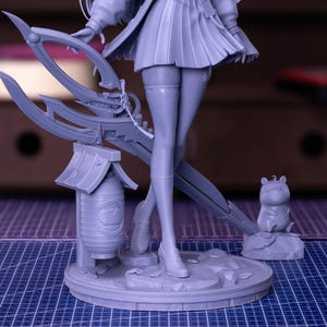 Puede incluir: Una figura gris de un personaje femenino con una falda plisada, sosteniendo un arma grande. La figura se encuentra sobre una base circular con una peque&ntilde;a linterna y una peque&ntilde;a estatua de animal. El fondo est&aacute; borroso.