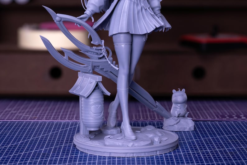 Puede incluir: Una figura gris de un personaje femenino con una falda plisada, sosteniendo un arma grande. La figura se encuentra sobre una base circular con una peque&ntilde;a linterna y una peque&ntilde;a estatua de animal. El fondo est&aacute; borroso.