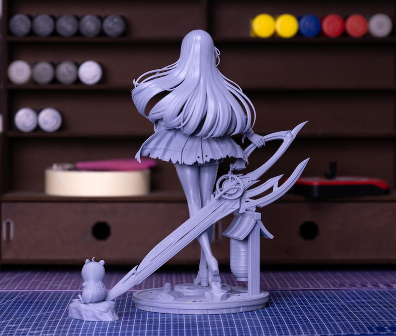 Puede incluir: Una figura gris sin pintar de un personaje femenino con cabello largo y suelto, sosteniendo un arma grande y ornamentada. La figura est&aacute; sobre una base circular, con una peque&ntilde;a figura de animal cerca. El fondo incluye una estanter&iacute;a de madera con materiales de pintura.