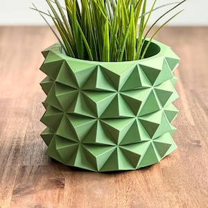 以下が含まれることがあります： 植物が入った緑色の幾何学的なプランター。プランターは質感のある素材で作られており、ユニークなデザインです。