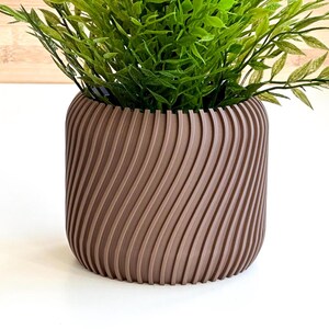 Pode incluir: Um vaso marrom com um design texturizado em espiral. O vaso está cheio de plantas verdes de folha.