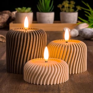 Op de afbeelding: Drie decoratieve kaarsen met een houtlook. De kaarsen zijn aangestoken en geven een warm licht. De kaarsen staan op een houten oppervlak.