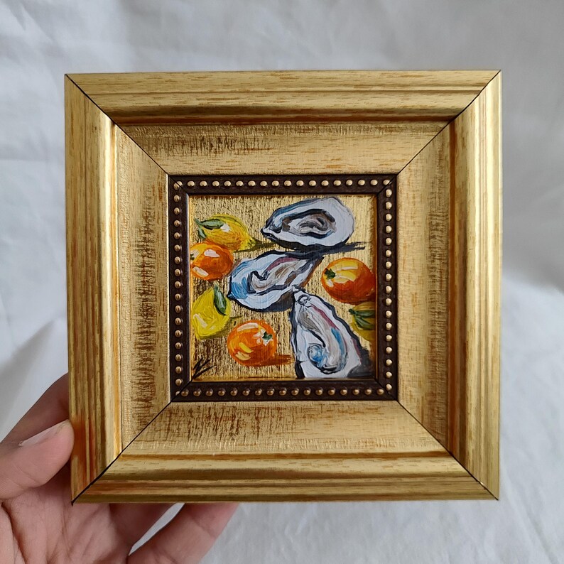 K&ouml;nnte beinhalten: Ein kleines, quadratisches Gem&auml;lde in einem goldenen Rahmen. Das Kunstwerk zeigt Austern und Zitrusfr&uuml;chte in Blau-, Orange- und Gelbt&ouml;nen. Der Rahmen hat eine strukturierte Oberfl&auml;che und einen dunkelbraunen Innenrand mit kleinen, dekorativen Punkten.