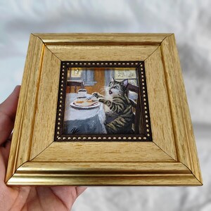 May include: A framed painting of a cat sitting at a table with a plate of food. The cat is looking at the food with a surprised expression. The painting is done in a realistic style and is set in a kitchen.