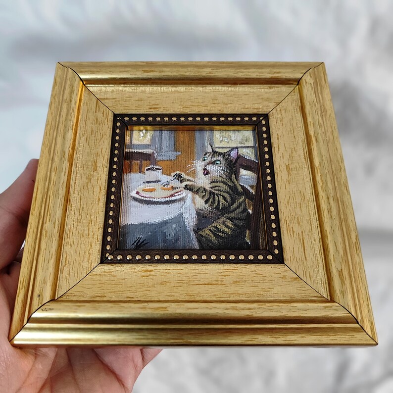 May include: A framed painting of a cat sitting at a table with a plate of food. The cat is looking at the food with a surprised expression. The painting is done in a realistic style and is set in a kitchen.