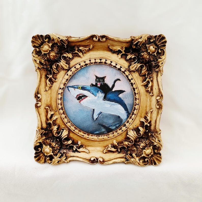 May include: A framed painting depicts a black cat riding a blue and white shark. The painting is set within a gold-colored, ornate frame. The artwork has a whimsical and playful theme.