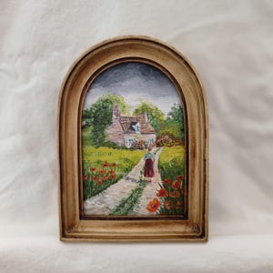 May include: A framed miniature painting depicts a cottage scene with a path leading to the house. The painting features a woman walking on the path, surrounded by red poppies and green grass. The frame is a light brown color.