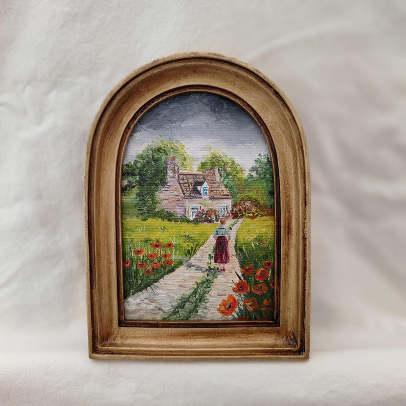 May include: A framed miniature painting depicts a cottage scene with a path leading to the house. The painting features a woman walking on the path, surrounded by red poppies and green grass. The frame is a light brown color.